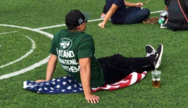 Man wearing "I Stand For The National Anthem" shirt sits on a field ... using the American Flag as his blanket.